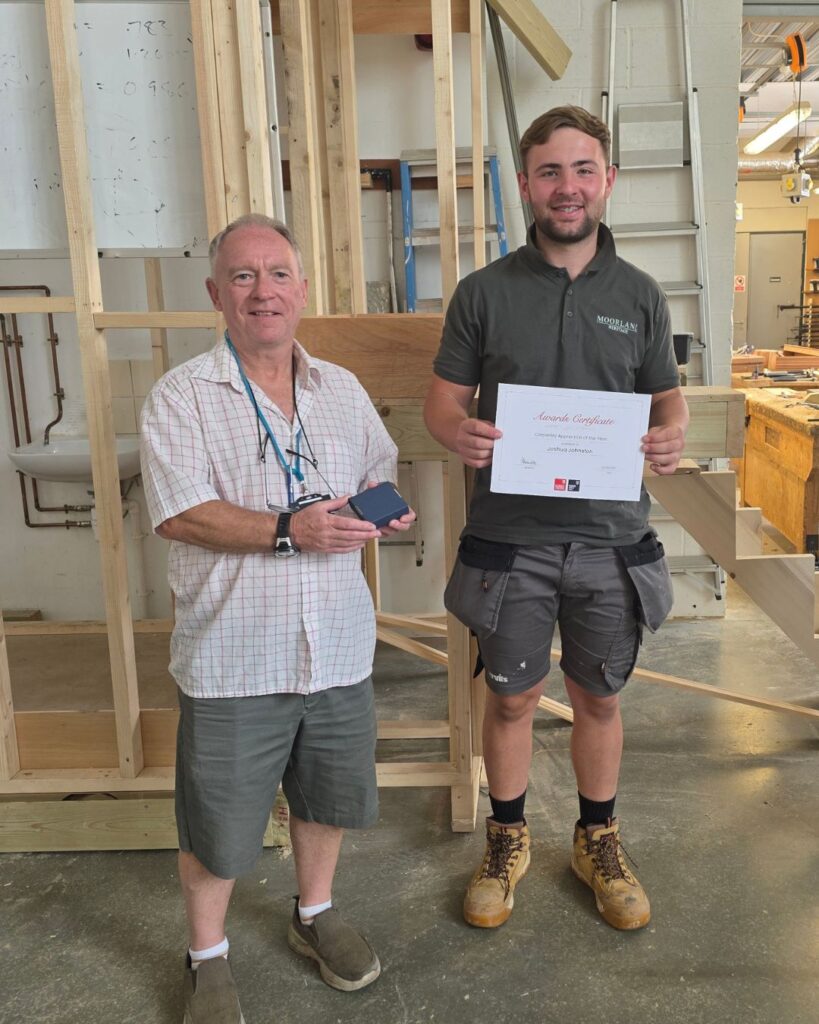Two men stood next to each other, with a certificate, in the carpentry workshop